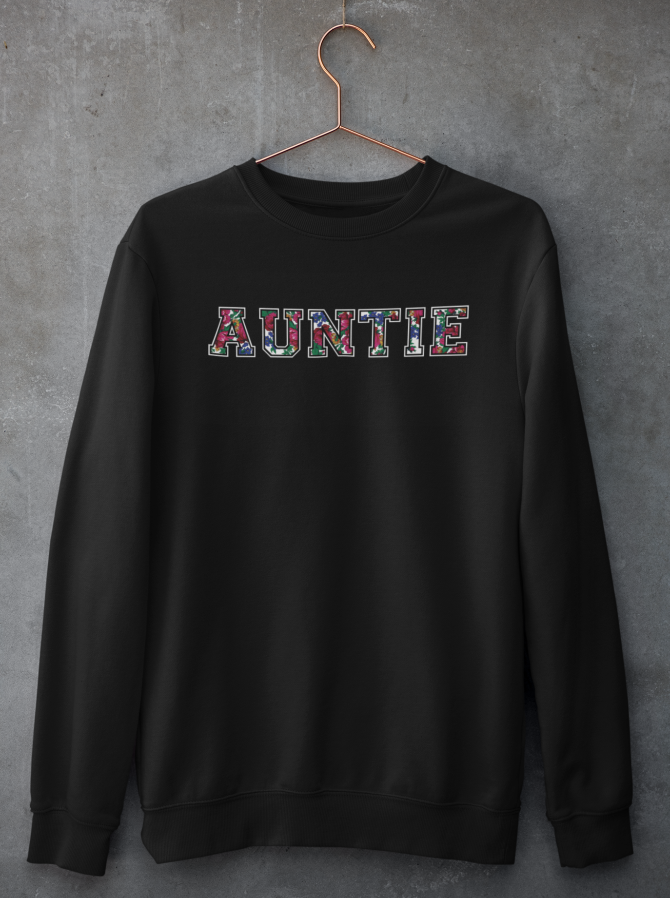 Black crewneck sweater with Auntie floral graphic on hanger against concrete backdrop